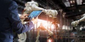 Persona che digita su un tablet circondata da braccia robotiche a lavoro dentro una fabbrica
