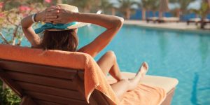 In primo piano donna sdraiata su un lettino a prendere il sole a bordo della piscina sullo sfondo